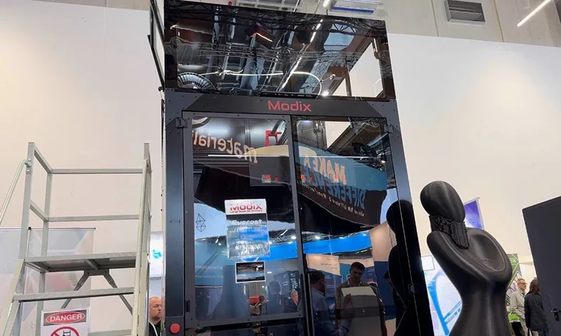 Modix Everest 3D printer with visible internal components and branding; includes a large sample part next to it in a tech exhibition setting.
