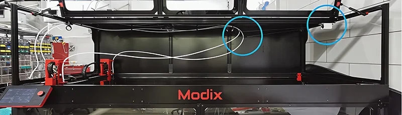 Modix Everest 3D printer in a workshop, featuring red accents and control panel, with filament samples illustrating its capabilities.
