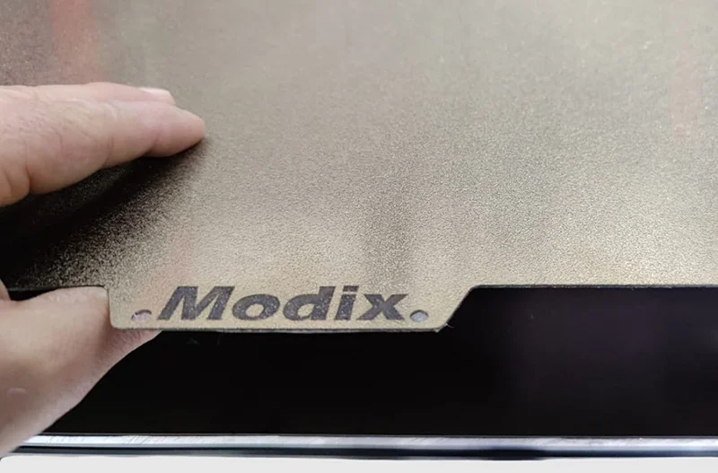 Hand lifting textured build plate labeled "Modix" on a 3D printer, showcasing material used for Modix Everest prints.