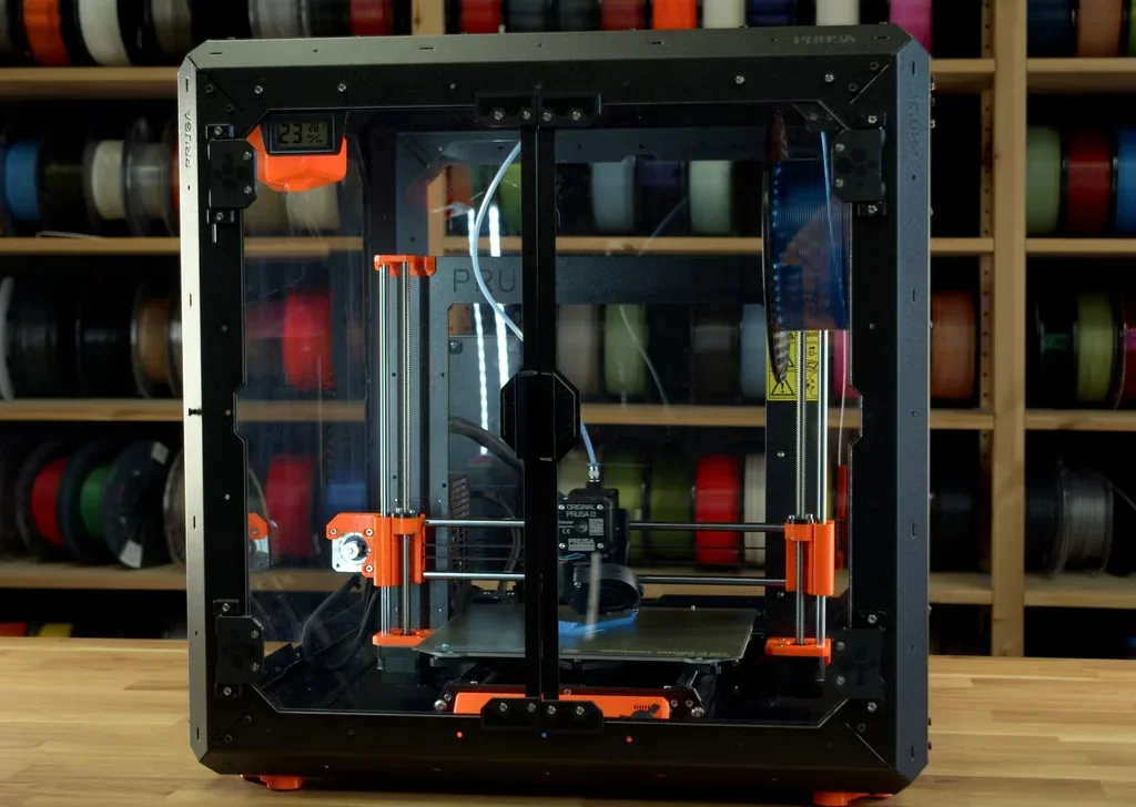 Original Prusa MK4S 3D printer kit with enclosed frame, orange accents, in a workshop with shelves of filament spools in the background.