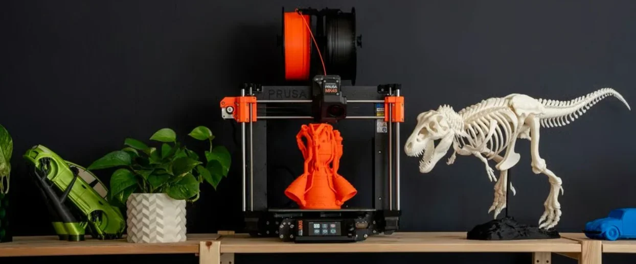Original Prusa MK4S 3D printer kit with orange filament, dinosaur skeleton, green model, and plant on a shelf.
