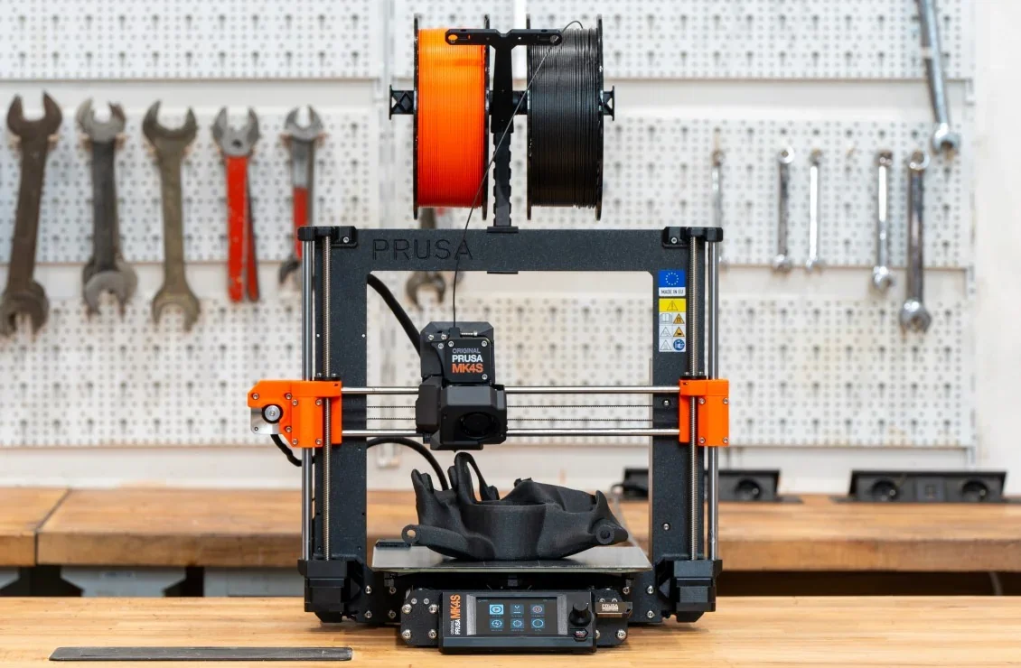 Original Prusa MK4S 3D printer kit in a workshop setting, featuring orange and black filament spools above, with tools on the wall in the background.