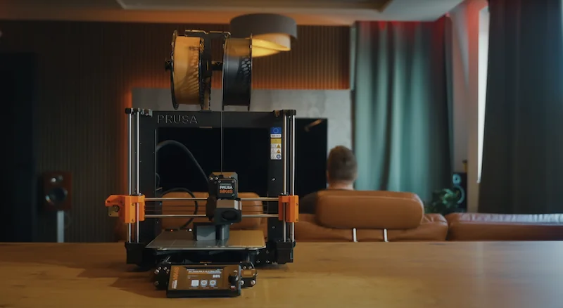 Original Prusa MK4S 3D printer kit on wooden table in modern room, with dual filament spools and control panel. Curved green curtains partly visible.