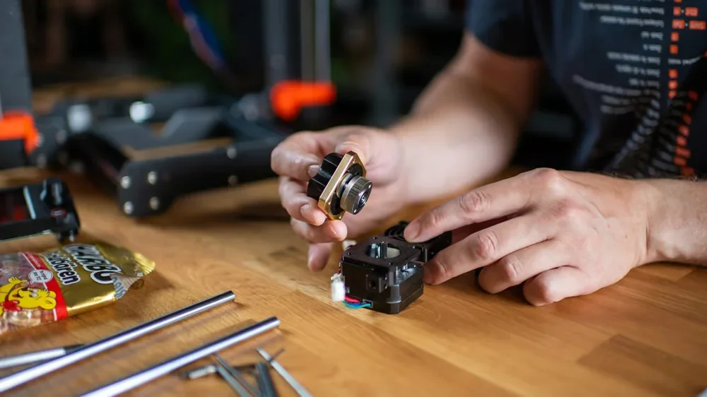 Hands assembling Original Prusa MK4S 3D printer kit parts on a wooden table with tools and snacks nearby.