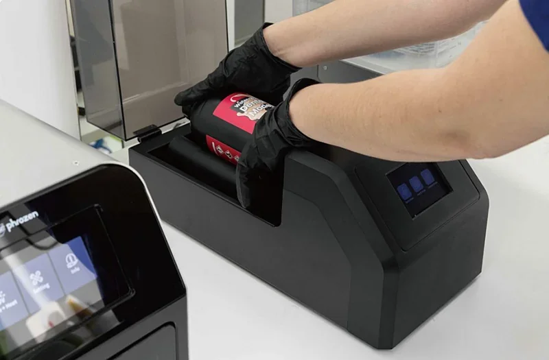 Phrozen Sonic CS+ 3D printer setup; hands in gloves insert red resin bottle into black base unit, displaying touchscreen panel.