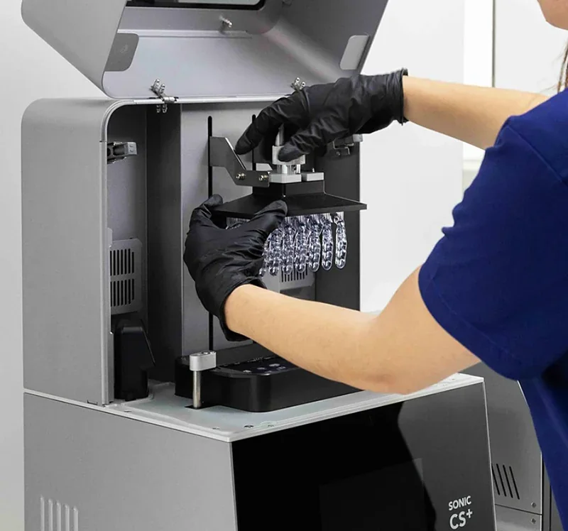 Person using Phrozen Sonic CS+ 3D printer with a resin model. Black gloves and open printer with parts displayed inside.