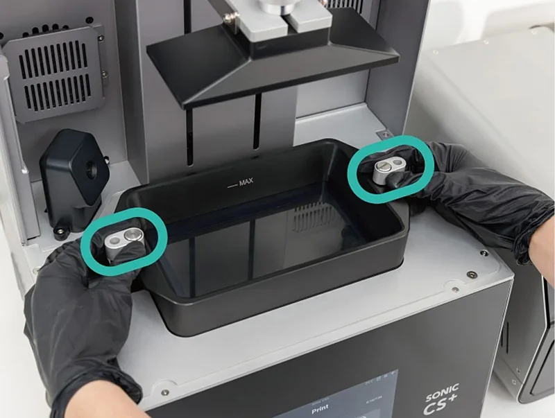 Phrozen Sonic CS+ printer open, gloved hands handling resin tray, two metal parts created by the printer highlighted, gray exterior, branding visible.