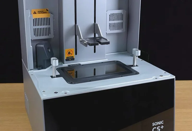 Phrozen Sonic CS+ 3D printer showing build area and platform. Gray panels with warning labels. Desktop setting, sleek and modern design.