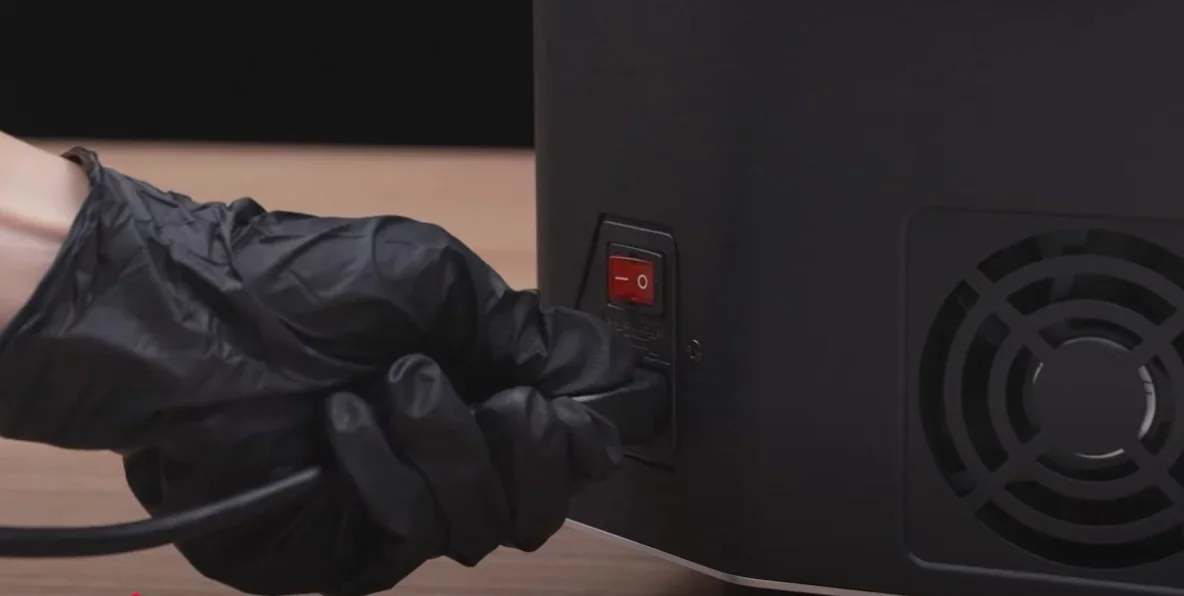 Plugging in the Phrozen Sonic Mega 8K 3D printer with a gloved hand next to a red power switch and fan on a wooden surface.