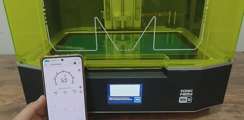 Phrozen Sonic Mega 8K 3D printer with green casing on a wooden table, a smartphone displays sound meter app, branding visible on the printer's front.