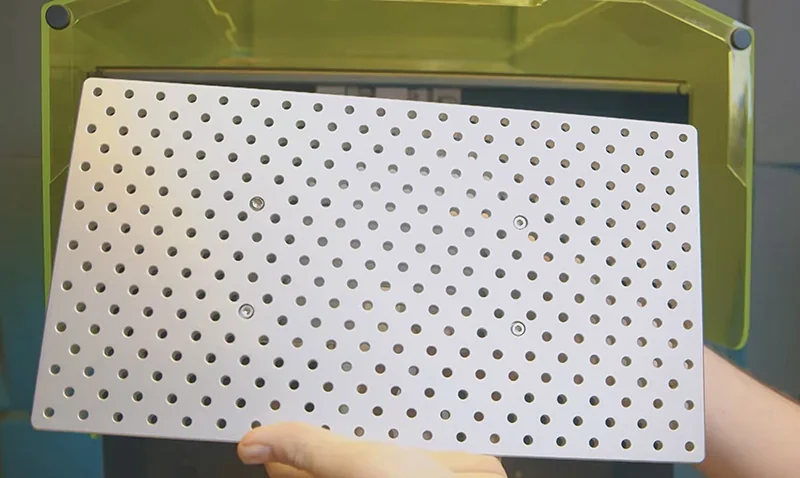 Hands hold perforated plate in front of Phrozen Sonic Mega 8K 3D printer, with clear green cover, used for printing high-resolution parts.