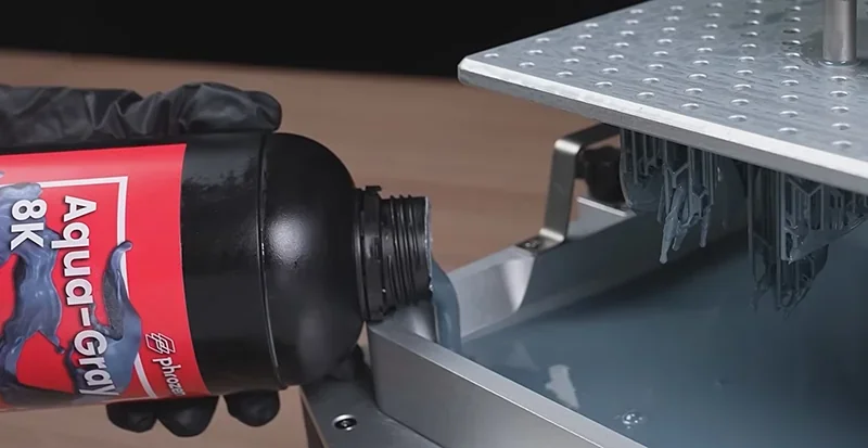 Phrozen Sonic Mega 8K pouring resin into a 3D printer vat with detailed 3D-printed parts visible, showcasing precision and high resolution.