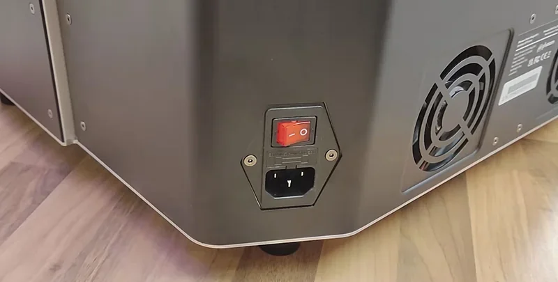 Phrozen Sonic Mega 8K power switch on a sleek, dark casing with visible fan vent, set on a wooden floor.