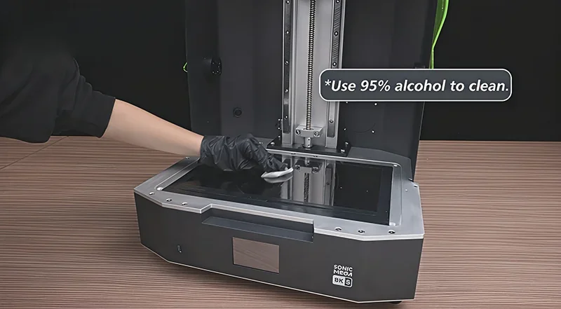 Phrozen Sonic Mega 8K 3D printer with open lid. Gloved hand cleans the print bed. Instruction reads: "Use 95% alcohol to clean." Visible branding present.