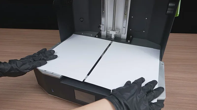 Phrozen Sonic Mega 8K 3D printer with open build area, hands in black gloves adjusting plates, on a wooden surface.