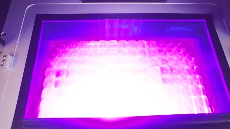 Phrozen Sonic Mega 8K V2 3D printer showing illuminated build plate with detailed resin print in progress, glowing purple light, machine enclosed setting.