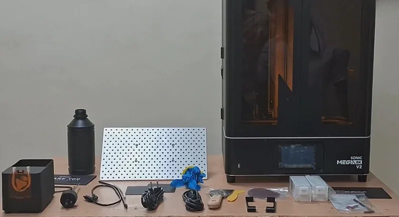 Phrozen Sonic Mega 8K V2 3D printer with resin bottle, perforated plate, cables, gloves, and accessories on a table.