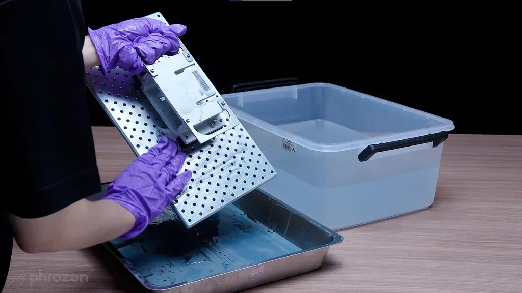 Person wearing purple gloves cleans Phrozen Sonic Mega 8K v2 printer plate over metal tray, next to a filled plastic container on wooden surface.