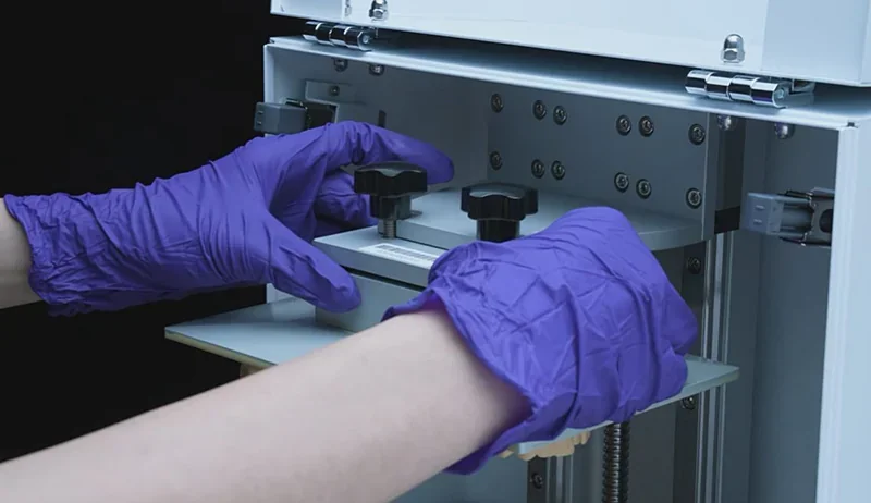 Hands in purple gloves adjusting Phrozen Sonic XL 4K Plus 3D printer bed, showcasing open panel and hardware, set in a workshop environment.