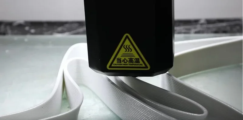 Piocreat G12 3D printer extruding white filament, creating a smooth, curved structure on a glass surface, warning label visible.