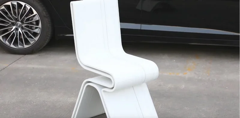 White chair on pavement, likely printed by Piocreat G12 3D printer, with sleek curves beside a car tire.