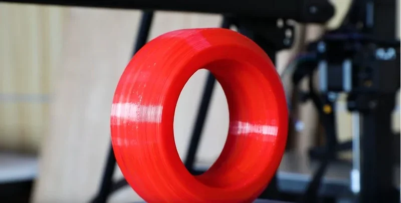 Red 3D-printed ring created by piocreat g5, with a blurred background of the printer in a workshop setting.