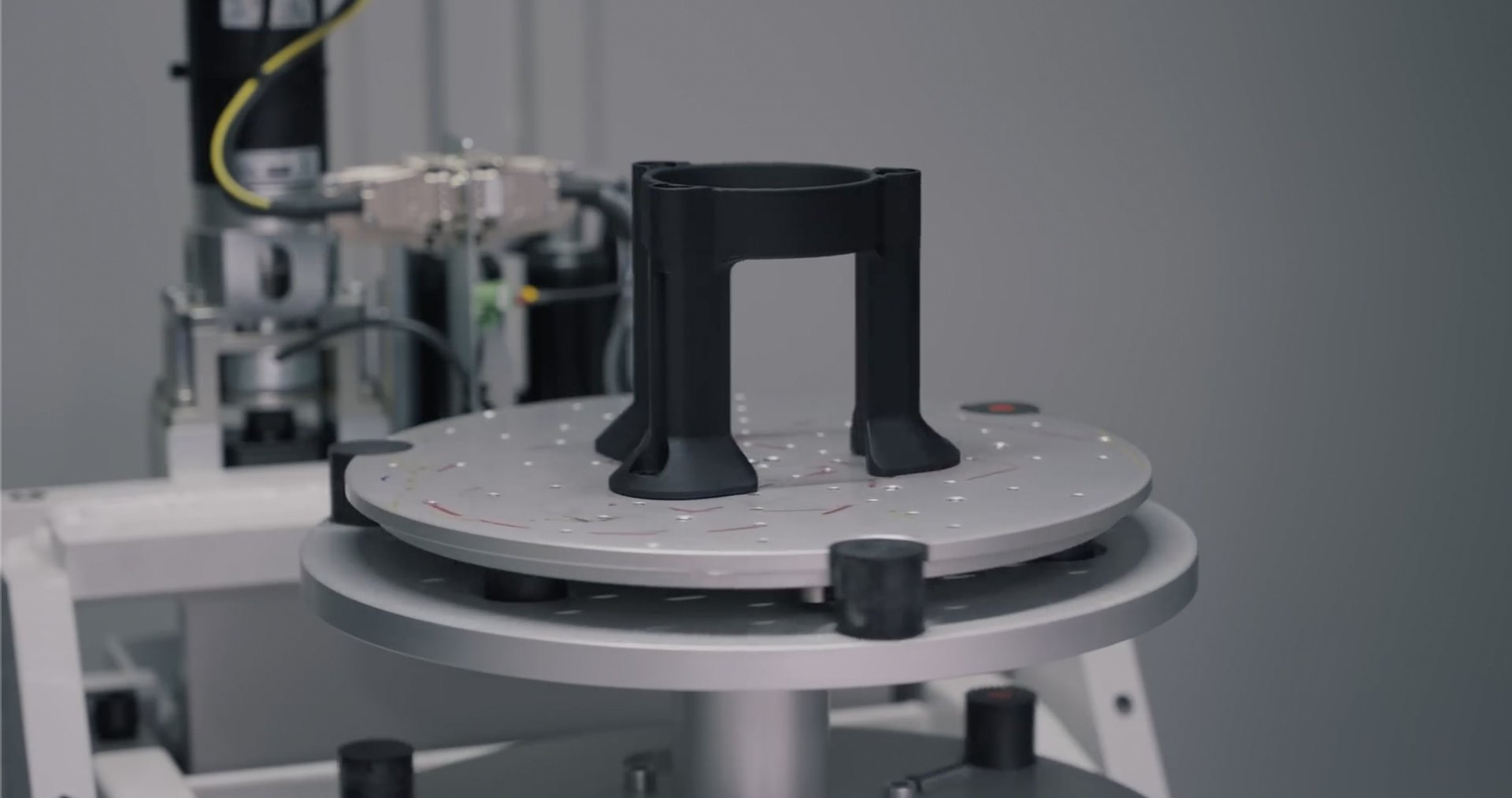 A cylindrical black 3D-printed object with four flared legs, sitting on a circular platform. The background shows parts of a 3D printer with cables and industrial components.