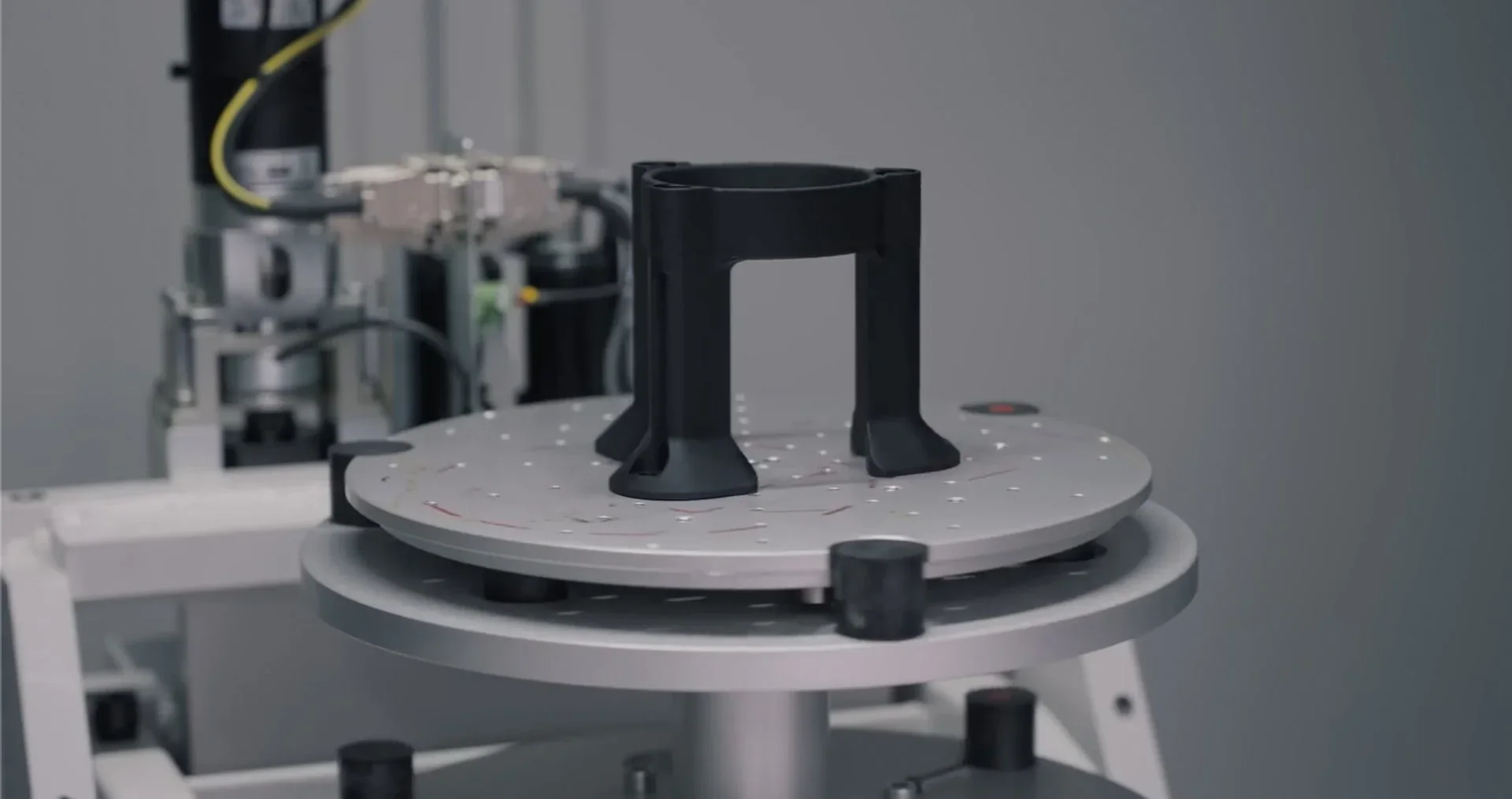 Prusa Core One 3D printer showcasing a black printed part on a rotating platform, surrounded by mechanical components in a technical setting.