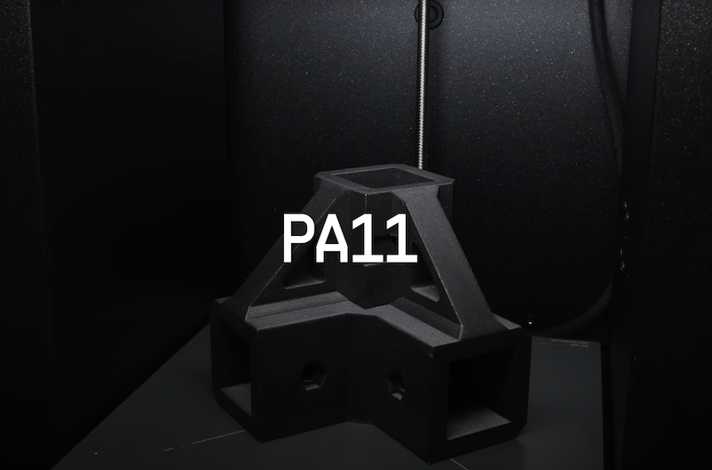 The image features a 3D printed geometric object with a truss-like structure, labeled "PA11," indicating it is made from polyamide 11 (Nylon 11). This sample print is placed inside