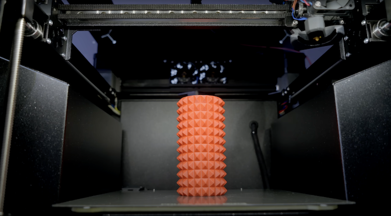 This image depicts the interior of a 3D printer with a red, cylindrical object on the print bed. The object has a spiked, textured surface, likely a print sample. The environment i