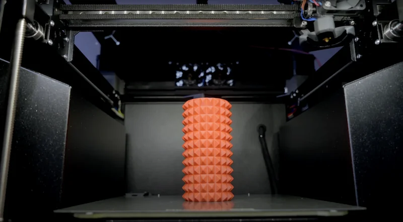 Prusa Core One 3D printer inside view with textured orange cylindrical object on the print bed, showcasing precise printing capabilities.
