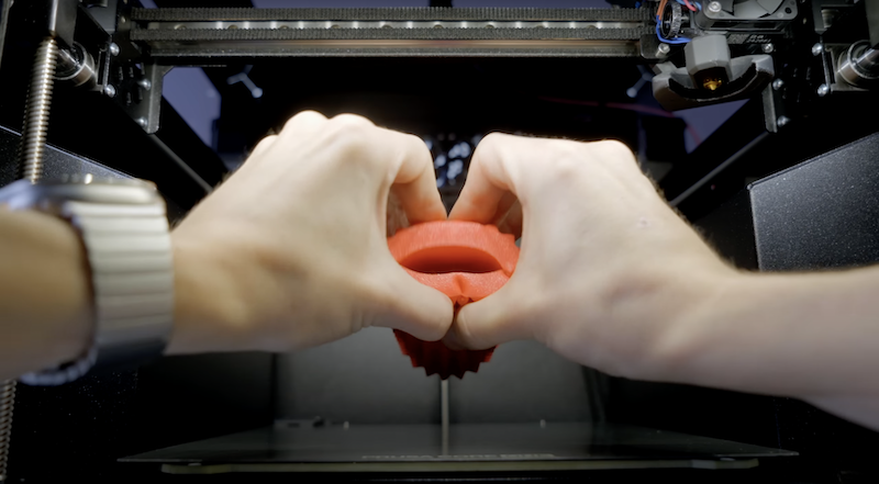 The interior of a 3D printer, likely the Prusa Core One, is shown with a person's hands carefully handling a flexible red 3D-printed part. The setup is compact, featuring advanced printer components such as linear rails and a sophisticated extruder, which may be characteristic of the Prusa's design. The lighting inside highlights the part being tested for its flexibility and resilience, which are key features of 3D-printed outputs.