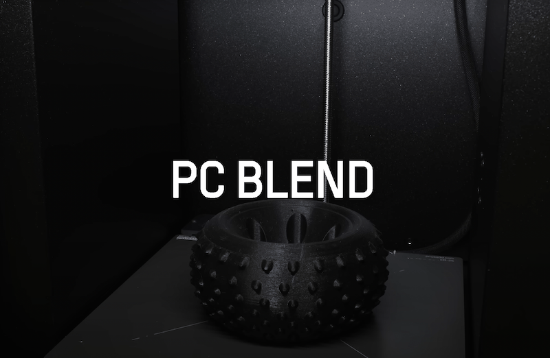 A 3D printed tire-like object is showcased on the print bed, labeled "PC BLEND." The object features a detailed, knobby tread pattern and was created with the Prusa Core One 3D Printer. Surrounding the item is the black, sleek enclosure of the printer, highlighting its manufacturing capabilities in a refined, professional setting.