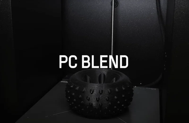 Prusa Core One 3D printer printing a black spiked wheel using PC Blend filament, showcased in a dark interior setting.