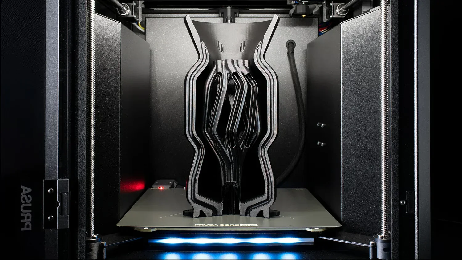 Prusa Core One 3D printer with a complex, abstract black structure on the print bed, surrounded by a sleek, modern frame and lit by LED lights.