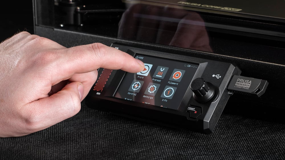A close-up of a 3D printer control panel. A hand is interacting with a touchscreen displaying various options, including "Print," "Preheat," "Filament," "Settings," and "Info." The