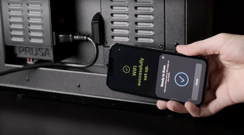 Prusa Core One 3D printer with phone showing successful Wi-Fi setup, highlighting tech connectivity.