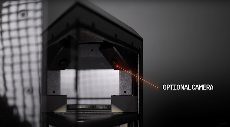 A close-up view of the Prusa Core One's upper section, highlighting an optional camera feature. A red laser pointer indicates the camera placement inside the black, angular frame. The focus is on the camera's integration into the printer’s structure, offering advanced monitoring capabilities. The dark, grid-patterned background emphasizes the technical sophistication of the design.