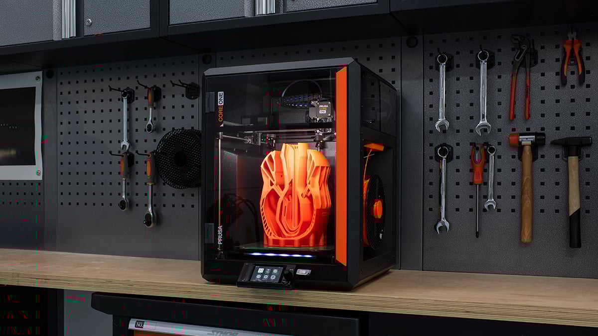 The image shows a 3D printer on a wooden workbench. The printer is enclosed in a black and orange casing, labeled "CORE ONE." Inside, a large, intricate orange 3D model is being pr