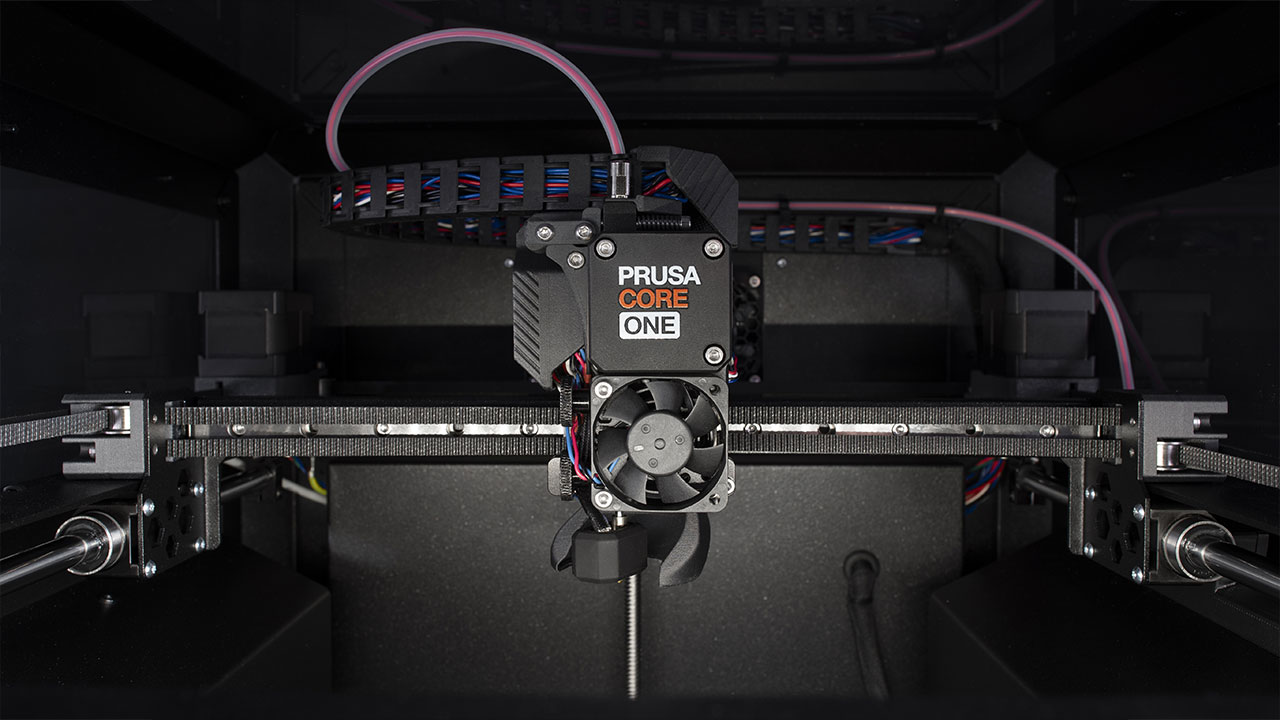 This image shows the internal components of a 3D printer, focusing on the print head labeled "PRUSA CORE ONE." The setup includes wires, belts, and a cooling fan. The print head is