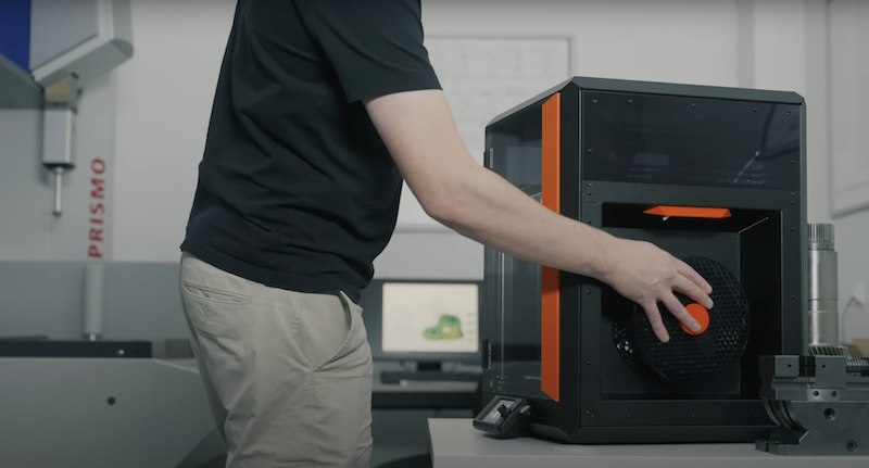 A person is interacting with a black and orange 3D printer. The printer has a transparent window, and there is a hand reaching inside, possibly adjusting or retrieving something. T
