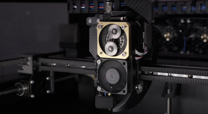 A close-up view of the Prusa Core One 3D Printer’s extrusion system reveals intricate technical components. Visible are the dual gears of the feeder mechanism, housed in a metallic frame, with a cooling fan attached to ensure optimal printing performance. The precision and integration of these components illustrate the advanced engineering and attention to detail in the printer’s design.