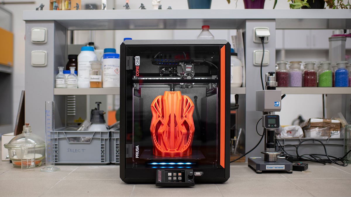 The image features a 3D printer in a laboratory or workshop. The printer is actively printing a large orange object with a complex, symmetrical design. The background is filled wit