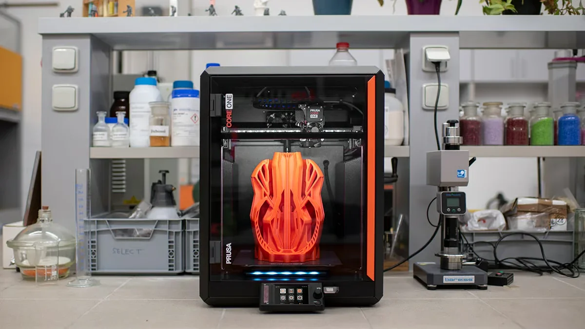 Prusa Core One 3D printer creating an intricate orange model in a lab setting, with shelves and equipment in the background.