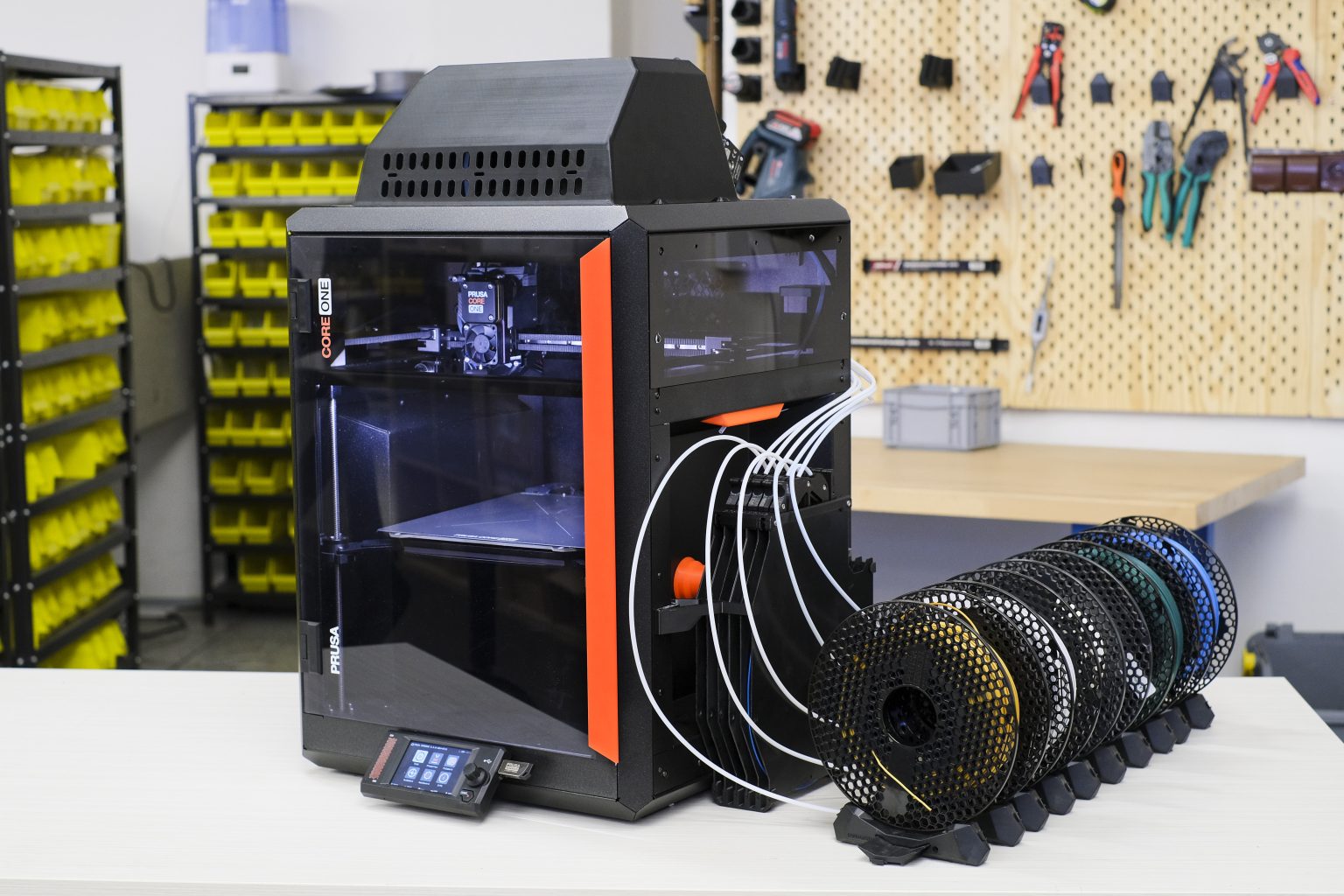 A 3D printer setup is shown in a workspace with a Prusa Core One printer prominently placed on a table. The printer is enclosed with a clear front panel, and multiple filament spoo
