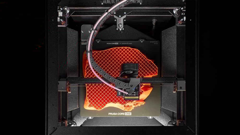 The interior view of a 3D printer actively creating an object. The print bed showcases a partially finished print with an orange lattice structure. The printer’s extruder and rails