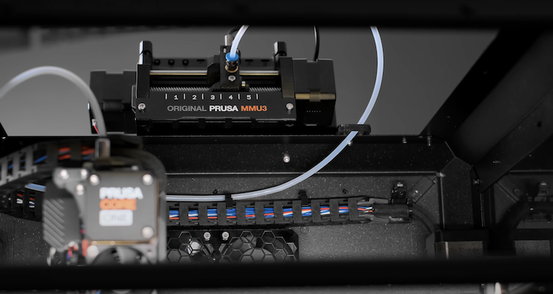 The image shows the interior of a 3D printer, focusing on the filament feeding mechanism. It features a component labeled "ORIGINAL PRUSA MMU3," with multiple slots for feeding dif