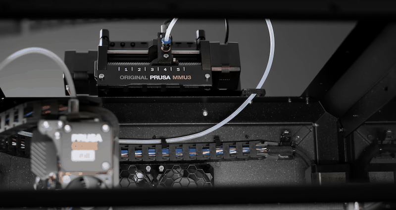 Close-up view of the Prusa Core One's internal mechanism featuring the Original Prusa MMU3 component. The image shows a detailed view of the filament feeder with five numbered slots for multicolor functionality. There is visible tubing connected to the feeder system, showcasing intricate wiring and component arrangement within the printer's frame.
