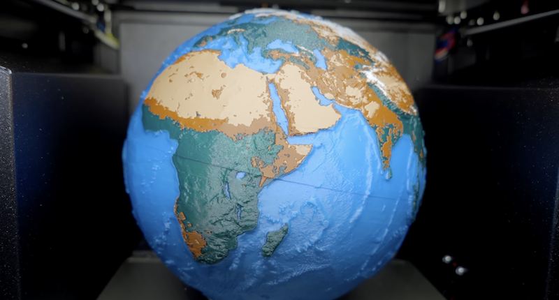 This image shows a 3D printer that has printed a detailed globe of Earth. The globe features realistic topography with various colors representing different elevations and terrains