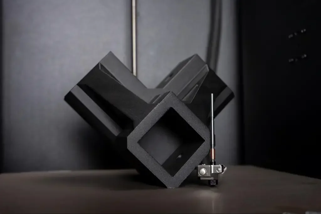 3D printed geometric part by Prusa Core One, black, angular design, with a metal component beside it, on a plain surface.